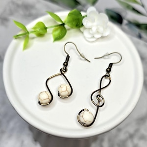 May include: A pair of handcrafted earrings featuring a musical note and treble clef design. The earrings are made with bronze-colored wire and adorned with white, marbled beads. The earrings are displayed on a white plate with greenery.