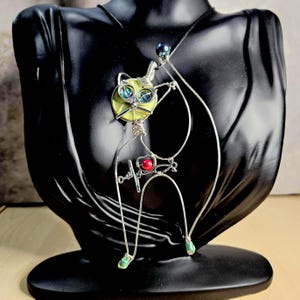 May include: A silver wire cat pendant necklace. The cat has a yellow face with blue eyes, a red heart, and green feet. The necklace is displayed on a black mannequin.