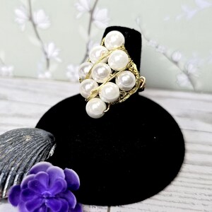 May include: A gold-toned wire ring featuring a cluster of white faux pearls. The pearls are arranged in a diamond shape and held together by the gold wire. The ring is displayed on a black velvet ring holder.
