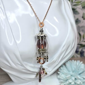 May include: A steampunk-style necklace with a copper chain and a pendant resembling a miniature cage. The cage holds a dark red orb and is adorned with silver and brass accents. The pendant has dangling beads and a small cluster of pink and gold beads.