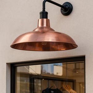 May include: A copper-colored, dome-shaped outdoor wall light fixture. The light has a black arm and mounting plate. The fixture is mounted on a light beige wall. The light is positioned above a window.