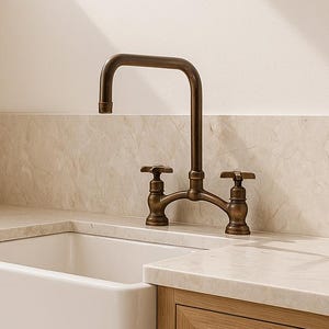 Puede incluir: Un fregadero de cocina con una pila de porcelana blanca y un grifo de bronce. El grifo tiene dos manijas y un caño curvo. El fregadero está empotrado en una encimera de color claro y gabinetes de madera.