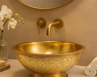 May include: A gold-colored, hammered metal sink with an ornate floral design. The sink is set on a marble countertop, with a matching faucet and mirror. The word "BRASSRY" is above the mirror.