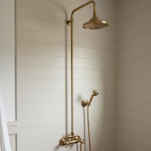 May include: A brass shower fixture with a round showerhead and a handheld showerhead. The showerhead is mounted on a long, vertical pipe. The fixture includes cross-shaped handles for water control. The shower is mounted on a white paneled wall.