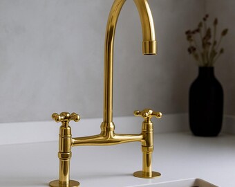 Puede incluir: Un grifo de cocina dorado con un caño curvo y dos manijas en forma de cruz. El grifo está montado sobre una encimera blanca. Un jarrón negro con flores secas está en el fondo.