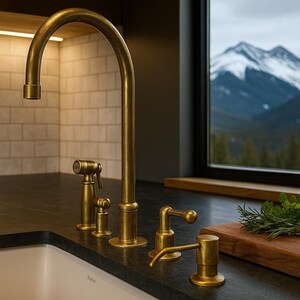 May include: A kitchen faucet set in a warm brass finish. The set includes a high-arc faucet, a side sprayer, and a soap dispenser. The faucet is set against a backdrop of white subway tile and a window with a mountain view.