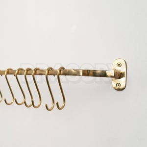 Könnte beinhalten: Eine messingfarbene Küchenreling mit mehreren S-förmigen Haken. Die Reling ist an einer weißen Wand befestigt, und die Haken sind gleichmäßig über die Stange verteilt. Das Design ist einfach und funktional, geeignet zum Aufhängen von Küchenutensilien.