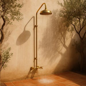 Puede incluir: Una ducha exterior de estilo vintage en latón cálido. El cabezal de la ducha es redondo, con agua cayendo. El accesorio está montado en una pared beige, con la sombra del cabezal y el accesorio proyectada sobre la pared. La ducha está rodeada de árboles.