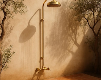 Puede incluir: Una ducha exterior de estilo vintage en latón cálido. El cabezal de la ducha es redondo, con agua cayendo. El accesorio está montado en una pared beige, con la sombra del cabezal y el accesorio proyectada sobre la pared. La ducha está rodeada de árboles.