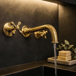 May include: A gold-colored wall-mounted faucet with cross-shaped handles. The faucet has a hammered texture and is positioned against a dark, textured wall. Water flows from the spout into a dark sink. Two bars of soap sit on a small tray.
