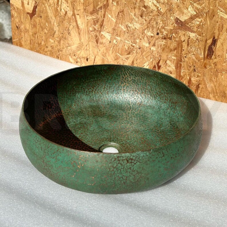May include: A green hammered copper vessel sink with a drain hole in the center. The sink has a distressed finish and a slightly rounded shape.