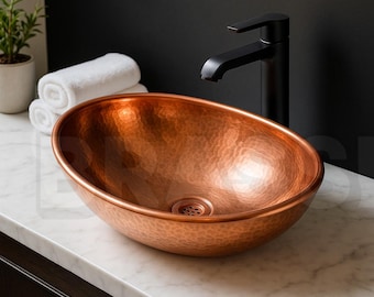 May include: Oval copper bathroom sink with a hammered texture. The sink is set on a white marble countertop, with a black faucet and a black wall in the background. Rolled white towels and a small potted plant are also visible.