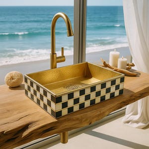Könnte beinhalten: Ein goldfarbenes rechteckiges Waschbecken mit einem schwarz-weißen Schachbrettmuster. Das Waschbecken hat einen goldenen Wasserhahn. Das Waschbecken steht auf einer Holzoberfläche mit Blick auf das Meer im Hintergrund. Dekorative Kerzen und ein Naturschwamm sind ebenfalls vorhanden.