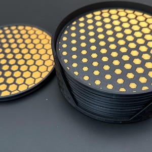 Könnte beinhalten: Set aus schwarzen Untersetzern mit einem gelb-schwarzen Wabenmuster. Die Untersetzer sind rund und werden mit einem passenden Halter geliefert. Das Design zeigt ein sich wiederholendes geometrisches Muster.