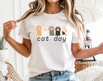 May include: White t-shirt with a colorful cat paw print design and the text "cat day" in black. The graphic features various cat paw prints in shades of orange, pink, gray, black, and tan. A casual, everyday tee.