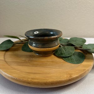 May include: A small, handcrafted ceramic bowl with a dark blue-green glaze. The bowl has a light brown band around the middle and sits on a round, wooden tray. Green leaves are arranged around the bowl.