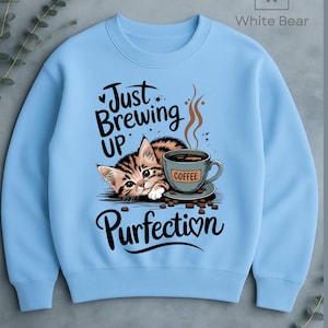 May include: Light blue sweatshirt featuring a tabby kitten with a coffee cup. The text reads "Just Brewing Up Purrfection" in a playful font. The coffee cup has the word "Coffee" on it. A cute and cozy design.