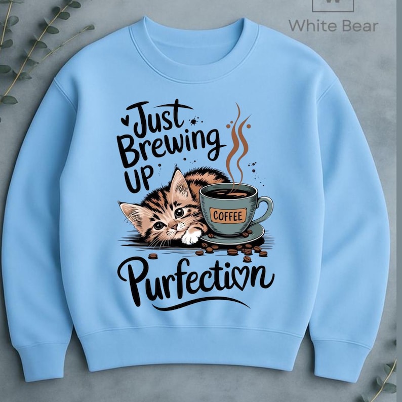 May include: Light blue sweatshirt featuring a tabby kitten with a coffee cup. The text reads "Just Brewing Up Purrfection" in a playful font. The coffee cup has the word "Coffee" on it. A cute and cozy design.