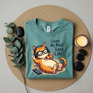 May include: A teal t-shirt featuring a cartoon orange tabby cat wearing headphones and glasses, holding a slice of pizza, and a remote control. The text "Living My Best Lazy Life!" is printed on the shirt. The shirt is folded on a woven mat.
