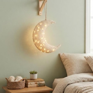 May include: A crescent moon-shaped wall lamp crafted from woven light-colored material, illuminated by small internal warm white LED lights. The lamp hangs from a light wood wall-mounted bracket by a thin cord, casting a soft glow in the room.