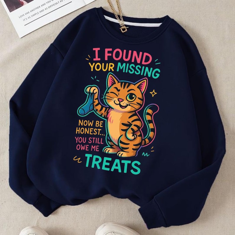 May include: Navy blue sweatshirt with a graphic of a winking orange tabby cat holding a blue sock. The text reads "I FOUND YOUR MISSING, NOW BE HONEST... YOU STILL OWE ME TREATS."