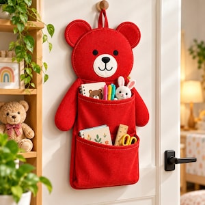 May include: A red felt wall organiser shaped like a bear with two pockets. The top pocket holds a small notebook, three markers, and a white plush rabbit. The bottom pocket contains a small notebook and a wooden ruler with yellow scissors.
