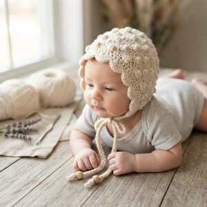 May include: A cream-colored crocheted bonnet featuring a textured bobble stitch pattern and a scalloped edge. The bonnet includes two long braided ties that hang down, each finished with a small, smooth, round wooden bead at the end.
