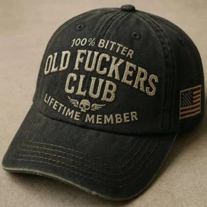 May include: A dark gray baseball cap with the text "100% Bitter Old Fuckers Club Lifetime Member" embroidered in off-white. Features a small American flag patch on the side. The cap has a curved brim and a distressed look.