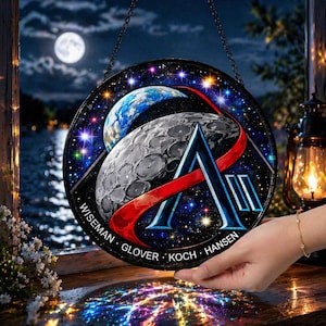 Könnte beinhalten: Rundes Glasfenster mit Weltraum-Motiv, das Erde, Mond und ein stilisiertes "A"-Logo zeigt. Es enthält einen roten Streifen, Sterne und die Namen "Wiseman, Glover, Koch, Hansen". Das Fenster wird von einer Kette gehalten.