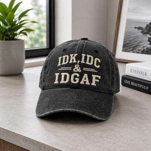 May include: A dark grey baseball cap featuring the embroidered text "IDK, IDC & IDGAF" in white. The cap has a distressed look and a curved peak. It is displayed on a grey surface.