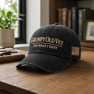 May include: Black baseball cap with the text "Grumpy Old Vet I do what I want" in gold lettering. The cap has a small American flag patch. The hat is on a wooden surface.