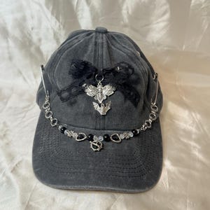 May include: A dark gray baseball cap adorned with a black lace bow, a silver moth pendant, and a beaded chain. The chain features black beads and decorative charms. The cap has a curved brim and a fabric strap closure.