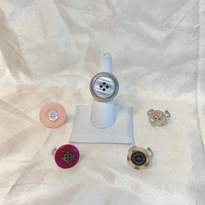 Puede incluir: Una colección de anillos con botones de varios colores y diseños. Un anillo presenta un botón grande e iridiscente con un engaste plateado. Otros anillos incluyen botones rosas, rojos y crema, cada uno con detalles y engastes únicos.