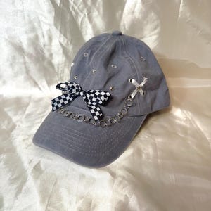 Puede incluir: Gorra de béisbol gris con un lazo a cuadros blanco y negro, una cadena plateada y detalles de estrellas y lazos. La gorra tiene una visera curva y está adornada con pequeños adornos plateados.