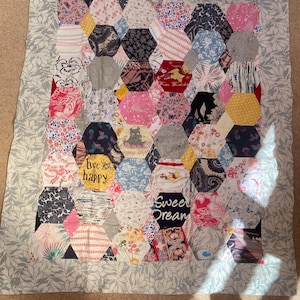 May include: A patchwork quilt made from a variety of fabrics in shades of pink, blue, grey, and yellow. The quilt is made up of hexagonal shapes and features phrases like "bee happy" and "sweet dream".