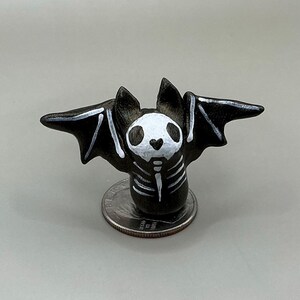 May include: A small, black bat figurine with white skeleton markings and a white skull-like face. The bat's wings are spread, and it is perched on a coin. The figurine is likely handmade and suitable for Halloween or gothic decor.