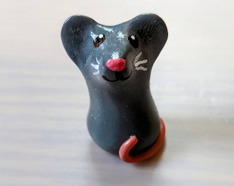May include: A small gray mouse figurine with a pink nose and white whiskers. The mouse is sitting up with its tail curled behind it.