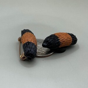 May include: Two small, detailed, black and orange-brown animal tail pins. One pin has a gold-coloured safety pin clasp attached. The pins are next to a silver coin on a light grey surface.