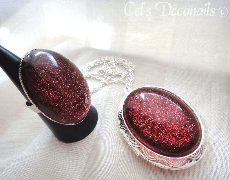 May include: A silver ring and a silver locket necklace with red glitter resin insets. The jewellery is on a white fabric background.