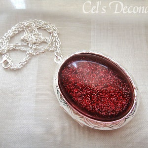 May include: A silver chain necklace with a silver oval locket pendant. The locket has a red glitter design inside.