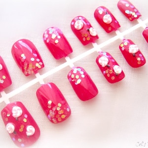 May include: A set of bright pink press-on nails. Some nails feature white flower designs and iridescent glitter accents. The nails are arranged on a white plastic holder, ready for application. The overall aesthetic is feminine and playful.