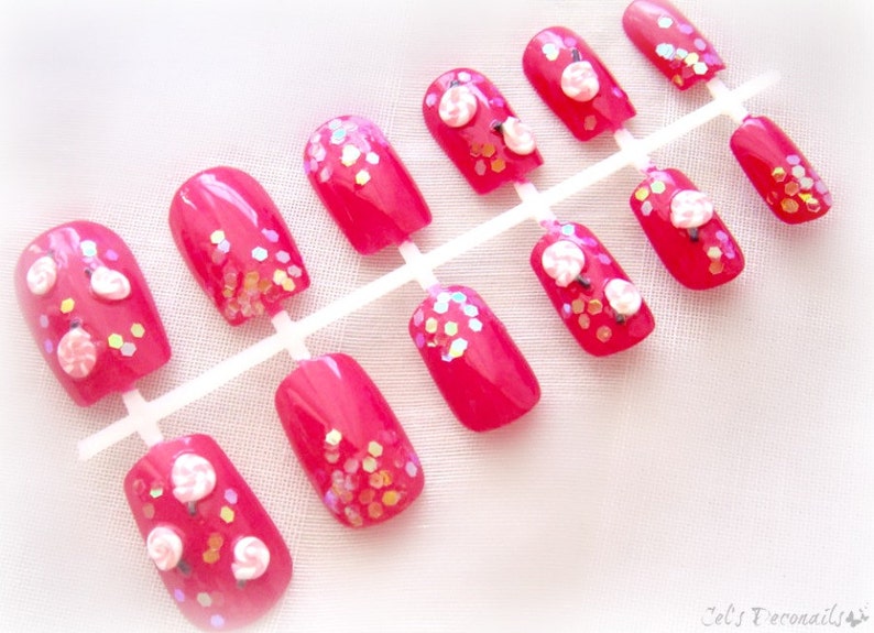 May include: A set of bright pink press-on nails. Some nails feature white flower designs and iridescent glitter accents. The nails are arranged on a white plastic holder, ready for application. The overall aesthetic is feminine and playful.