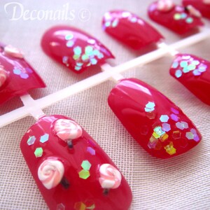 May include: Red artificial nails with glitter and candy decorations. The nails are arranged on a white plastic holder. The text "Cel's Deconails" is visible in the upper left corner.