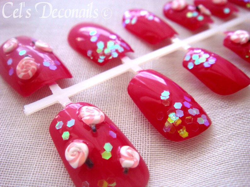 May include: Red artificial nails with glitter and candy decorations. The nails are arranged on a white plastic holder. The text "Cel's Deconails" is visible in the upper left corner.