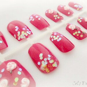May include: A set of press-on nails in a vibrant pink hue. Some nails feature iridescent glitter accents, while others are adorned with small, delicate flower designs. The nails are arranged on a white plastic backing.
