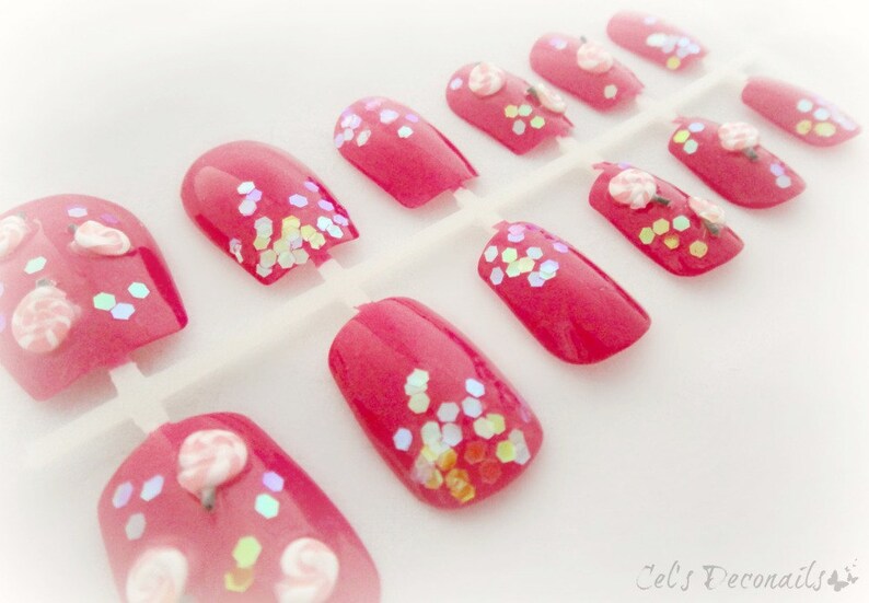 May include: A set of press-on nails in a vibrant pink hue. Some nails feature iridescent glitter accents, while others are adorned with small, delicate flower designs. The nails are arranged on a white plastic backing.
