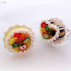 Könnte beinhalten: Zwei Miniatur-Kuchen mit Obstbelag, einer mit gelber Basis und weißem Frosting, der andere mit schwarz-weiß gestreiftem Boden und weißem Frosting. Beide Kuchen sind mit verschiedenen bunten Obstscheiben belegt.