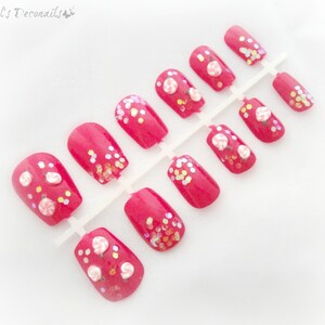 May include: A set of bright pink press-on nails. Some nails feature small, white and pink flower designs, while others have iridescent glitter accents. The nails are arranged on a white plastic holder, ready for application.