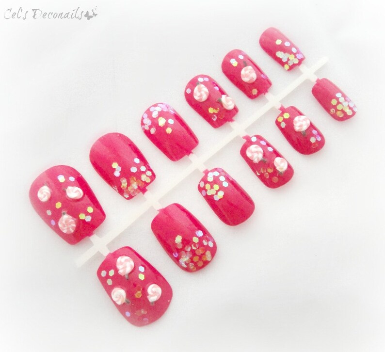 May include: A set of bright pink press-on nails. Some nails feature small, white and pink flower designs, while others have iridescent glitter accents. The nails are arranged on a white plastic holder, ready for application.