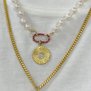 May include: A layered necklace set featuring a strand of white pearls connected to a gold-tone oval clasp with pink stones and a round gold pendant with the letter S, and a gold-tone chain with a heart-shaped pendant featuring a star design.